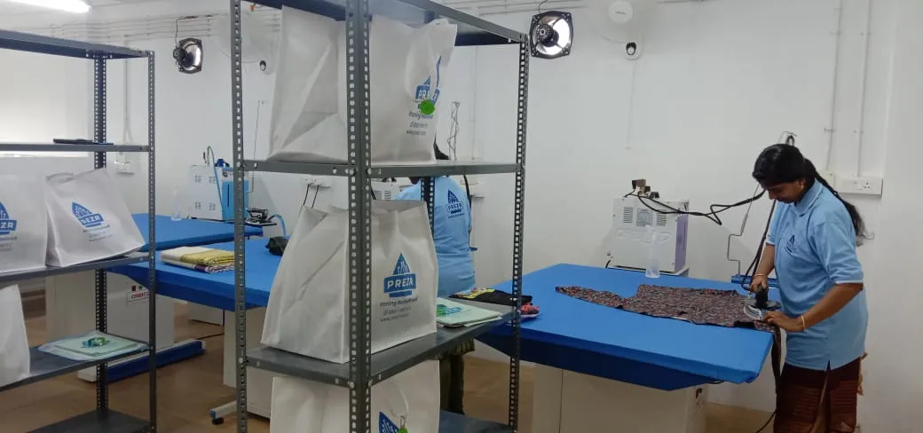 A member of the Prezr team steam pressing a patterned garment on a blue-topped table, with finished orders in branded bags on the shelves behind.