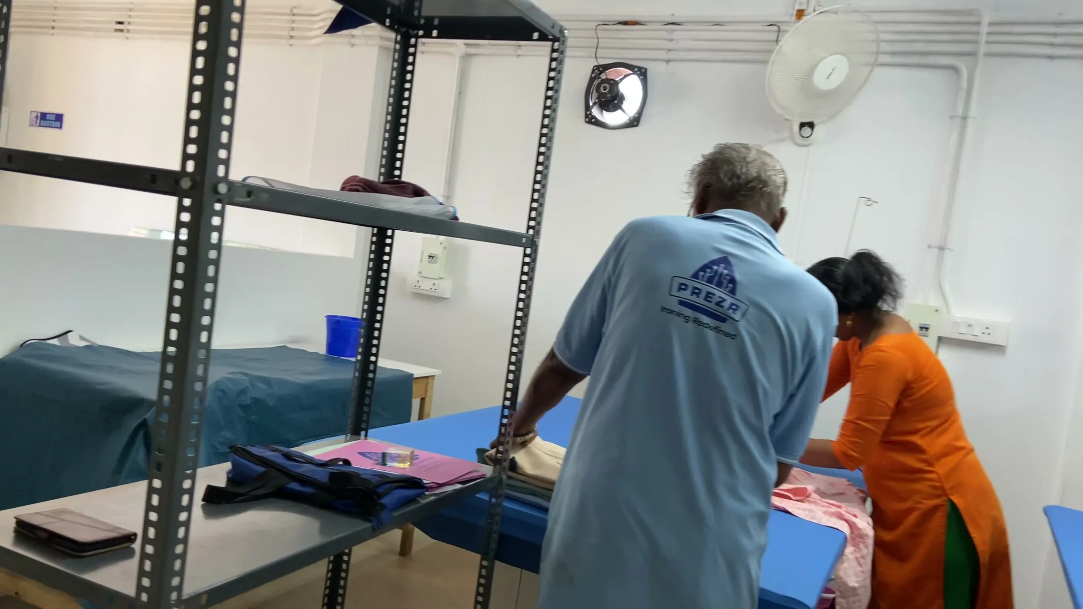 Two members of the Prezr workshop team working at the pressing tables, one in a Prezr shirt reading Ironing Redefined, with finished orders stacked on the racks beside them.